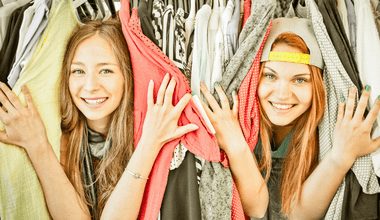 Two young white female teenagers - possibly twins - poking their heads out of clothing hanging in a closet. One of the girls is wearing a backward baseball cap.