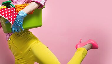 Cropped photo taken against a pink background of a woman in tight, bright yellow jeans and hot pink high heels. She's wearig bright blue rubber gloves to do her spring clearing and cleaning, and carrying a green cleaning bucket with a red polka-dot cleaning rag.