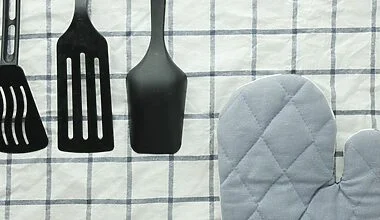 Photograph of three black silicone cooking utensils and an oven mitt on a checked tablecloth, some of the kitchen essentials for a first apartment.