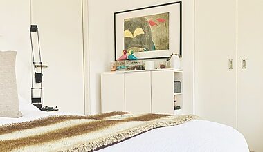 Photo of a small, uncluttered, tidy bedroom with no closet, for a post about no closet bedroom ideas. The walls and door are white, and there is a white high board from Ligne Roset upon which clear acrylic jewelery boxes from Muji are kept. A colorful painting hangs on the white wall. The bedspread is white, and the bed has a beige pillow and fur runner on it.