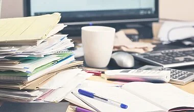 Photo of a messy desk piled high with files, notebooks, random paperwork, a coffee mug and a glass, pens, calculator, and a mobile handset. Everything is strewn around in front of the computer monitor and keyboard and looks very cluttered and shows how difficult it can be to organise a desk without drawers.