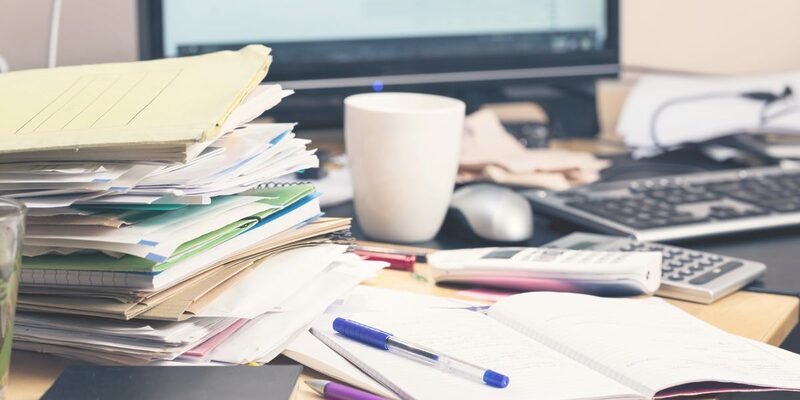 Photo of a messy desk piled high with files, notebooks, random paperwork, a coffee mug and a glass, pens, calculator, and a mobile handset. Everything is strewn around in front of the computer monitor and keyboard and looks very cluttered and shows how difficult it can be to organise a desk without drawers.