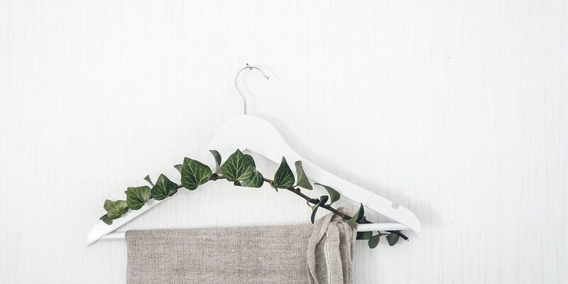 Photo of a beige linen pair of trousers hanging from a white hanger with ivy draped over it, in front of a white wall for an article titled why choose sustainable fashion.