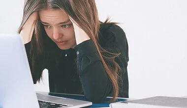 What is mental clutter? Photo of a young woman with long brown hair hunched over her laptop, cradling her head in her hands and looking very worried.