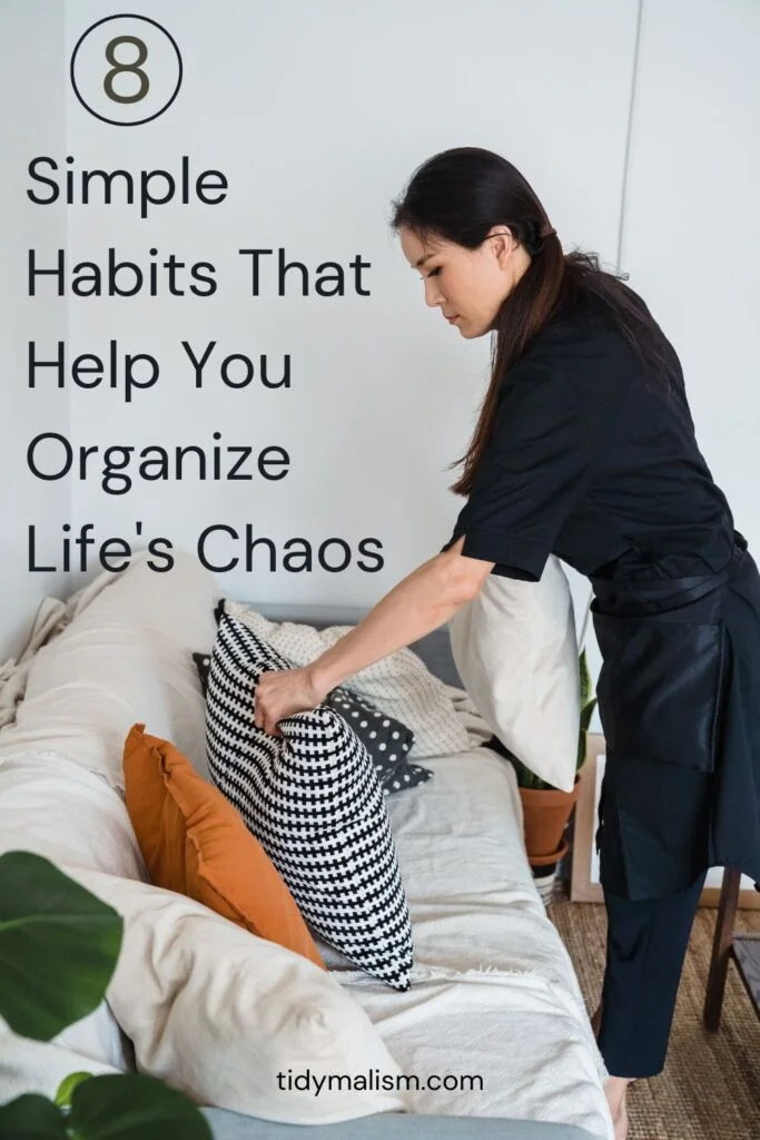 Asian woman with long dark hair pulled back, wearing an all black outfit and black apron, tidying up the pillows on her cream colored couch as part of her tidying routine in how to organise a chaotic life.