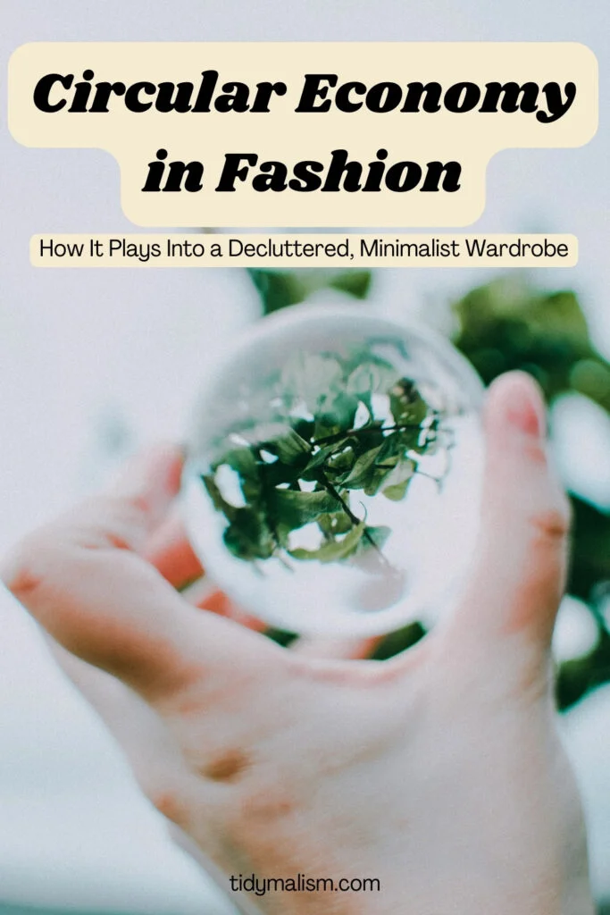 White male hand holding a crystal ball though which we can see green leaves and branches. Caption reads circular economy in fashion: how it plays into a decluttered, minimalist wardrobe.