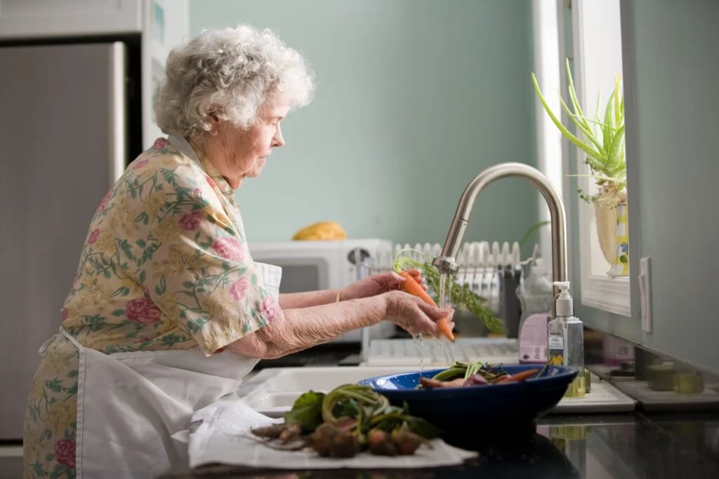 Elderly woman in a floral dress and white apron standing at her kitchen sink and washing carrots.