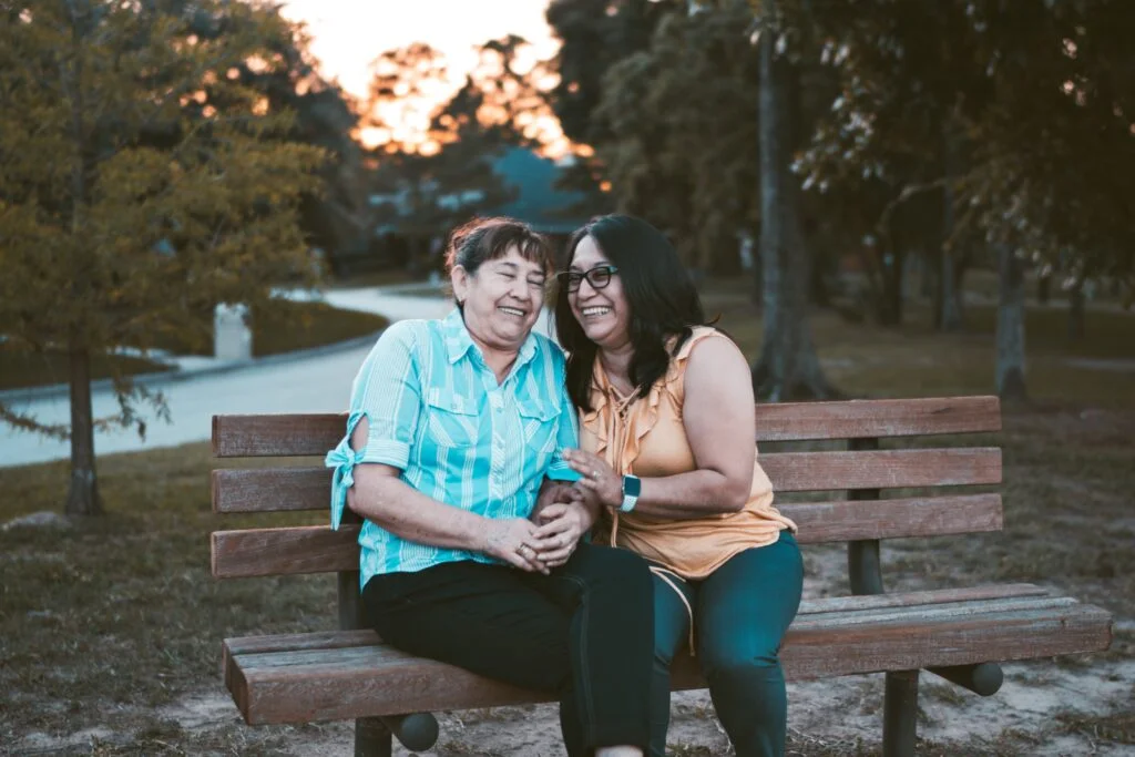 Two middle-aged woman sitting closely on a park bench and having a laugh with each other.