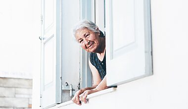 Elderly woman with white hair leaning out of her window and looking directly in the camera with a friendly smile on her face.