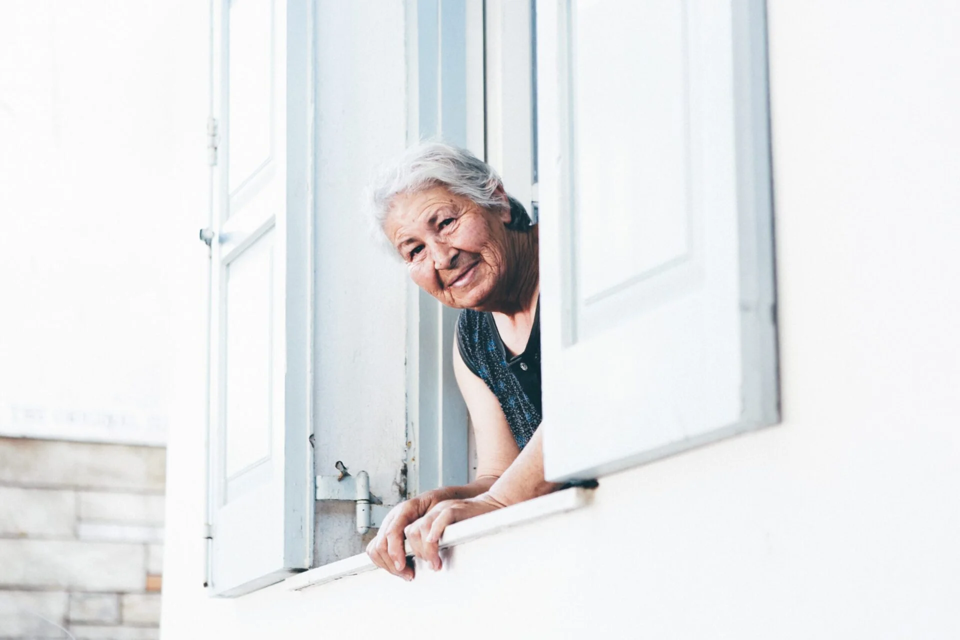 Elderly woman with white hair leaning out of her window and looking directly in the camera with a friendly smile on her face.