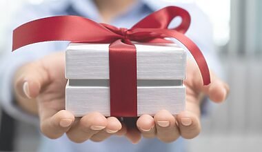 Photo of two outstretched hands holding a small white, minimalist gift box wrapped in a red ribbon.