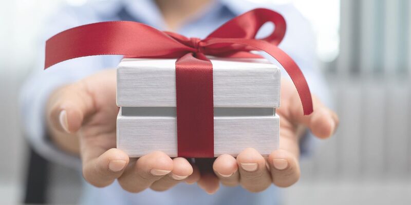 Photo of two outstretched hands holding a small white, minimalist gift box wrapped in a red ribbon.