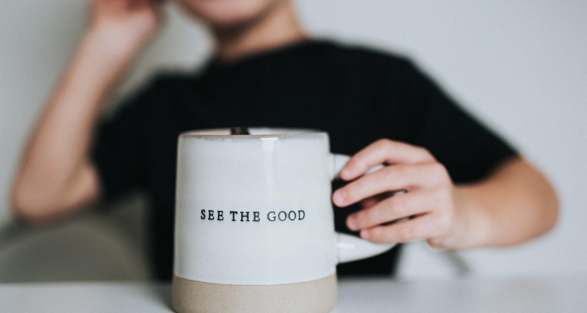 Cropped photo by Nathan Dumlao of a young person sitting at a table, with hand on a mug that has a positivity message "see the good."