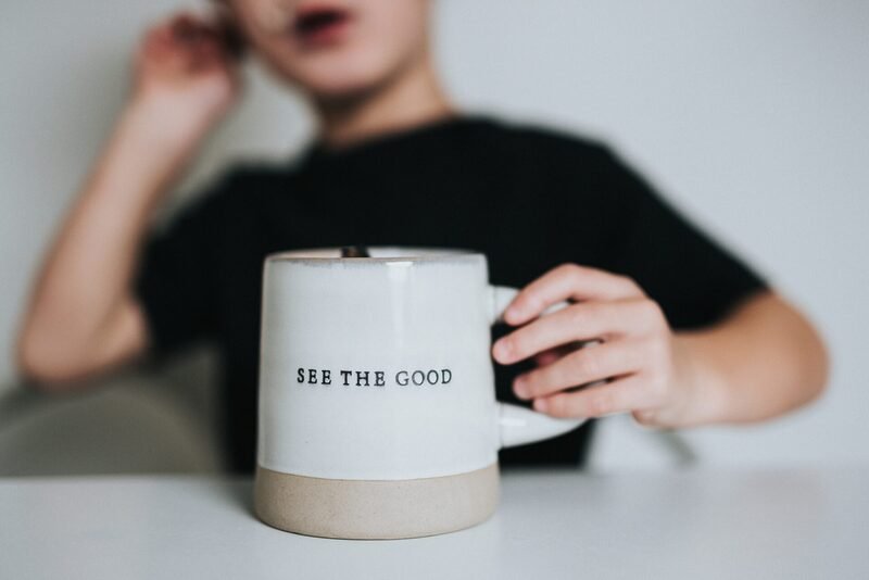 Cropped photo by Nathan Dumlao of a young person sitting at a table, with hand on a mug that has a positivity message "see the good."