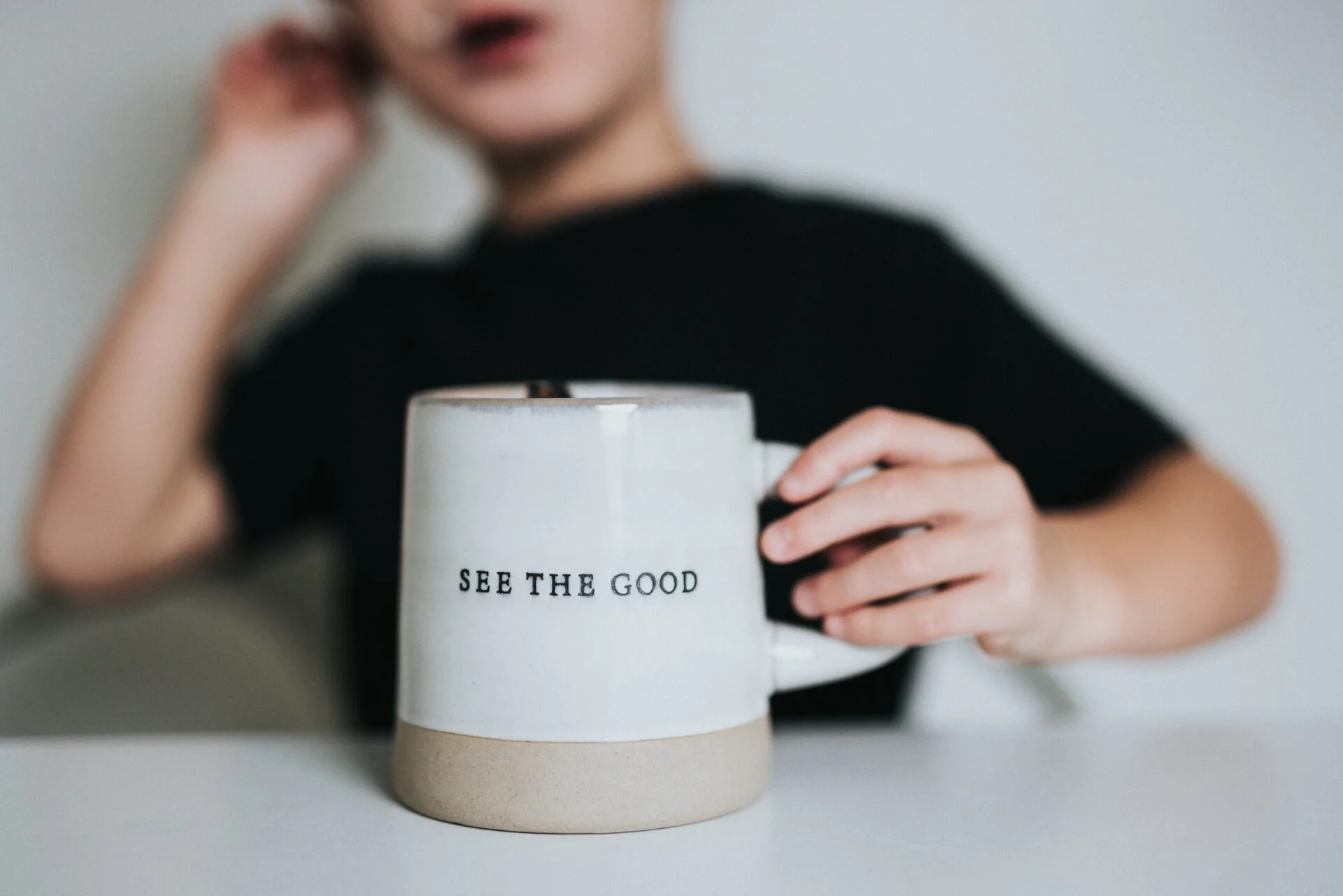Cropped photo by Nathan Dumlao of a young person sitting at a table, with hand on a mug that has a positivity message "see the good."