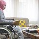 Young woman in a hijab, sitting in a wheelchair in her living room and tidying things up on the coffee table.