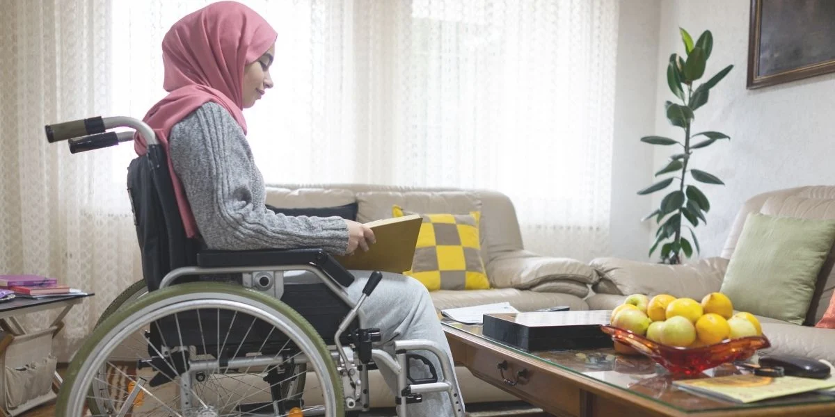 Young woman in a hijab, sitting in a wheelchair in her living room and tidying things up on the coffee table.