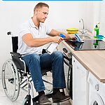 Young man with white skin and blond hair sitting in a wheelchair at his kitchen stove, wiping it down with a bottle of spray cleanser and soft rag.