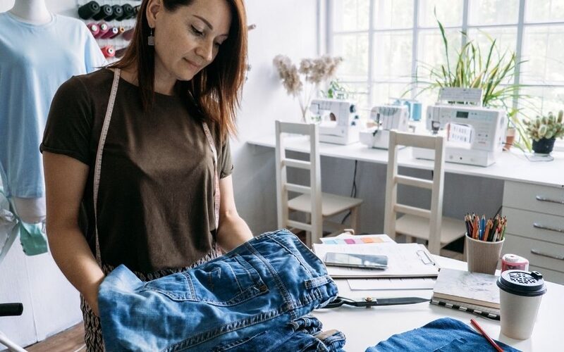 A white woman with shoulder-length brown hair standing at a large white workspace with a pile of denim jeans. Behind her are sewing machines. She is repairing and upcycling the old jeans, which have a long lifespan.
