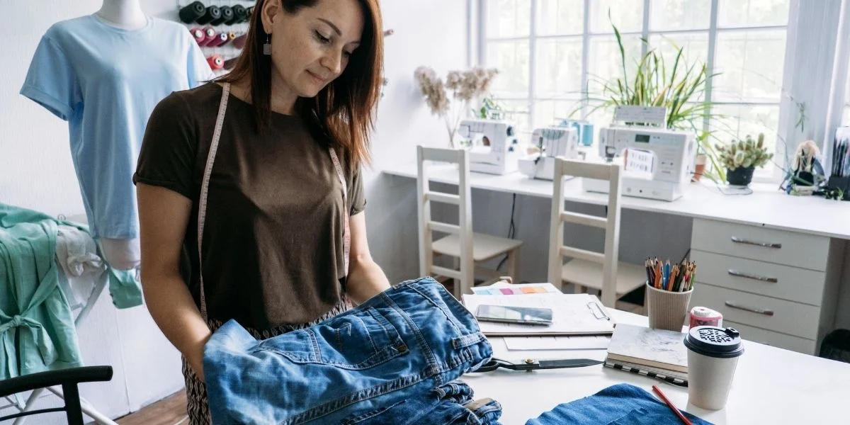 woman with shoulder-length brown hair standing at a large white workspace with a pile of denim jeans. Behind her are sewing machines. She is repairing and upcycling the old jeans, which have a long lifespan.