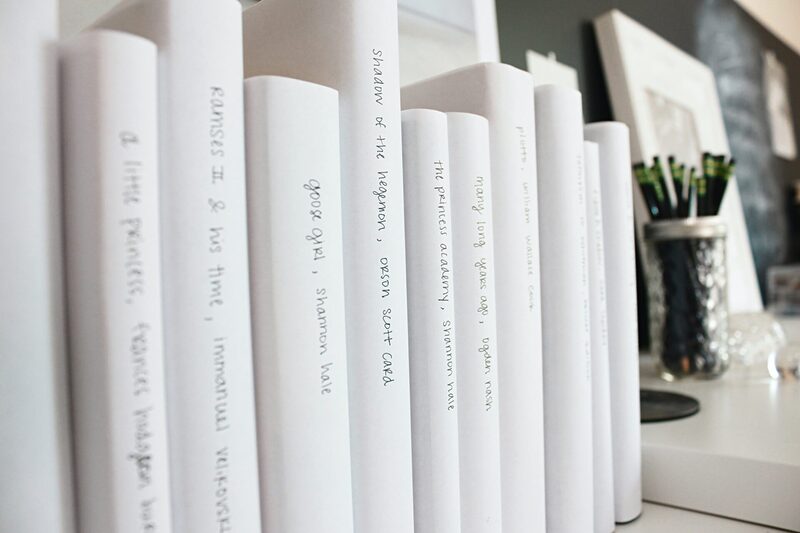 Close-up photograph of a bookshelf showing books all in white dust jackets with neat titles hand written in black pen.