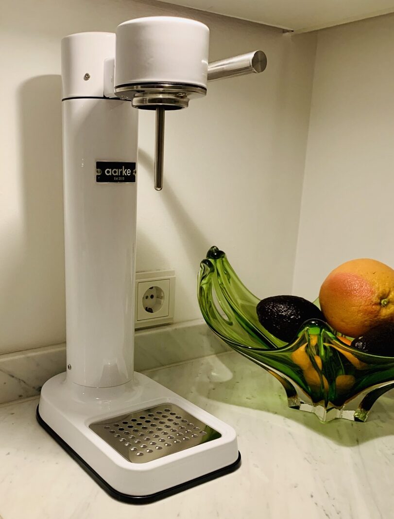 Aarke carbonator in white, on a white marble kitchen countertop to the left of a green Murano glass fruit bowl with oranges and avocados in it.